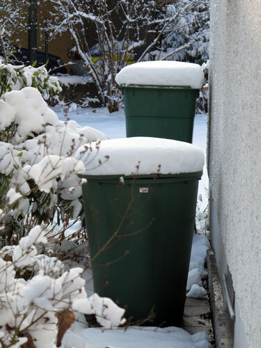 Two green water butts topped with snow by the house wall in winter