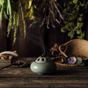 Smoking ceramic incense burner on a wooden table, with bunches of dried herbs behind