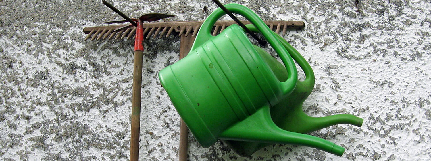 Two green watering cans and a rake hanging on a house wall